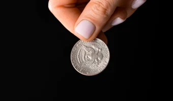Mână ținând o monedă argintie între degete, pe fundal negru